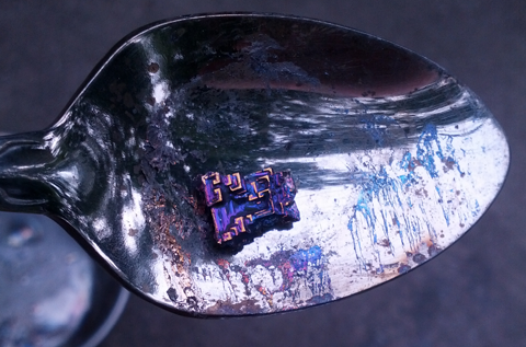 A bismuth crystal sits in a kitchen spoon, displaying iridescent purple and orange coloration.