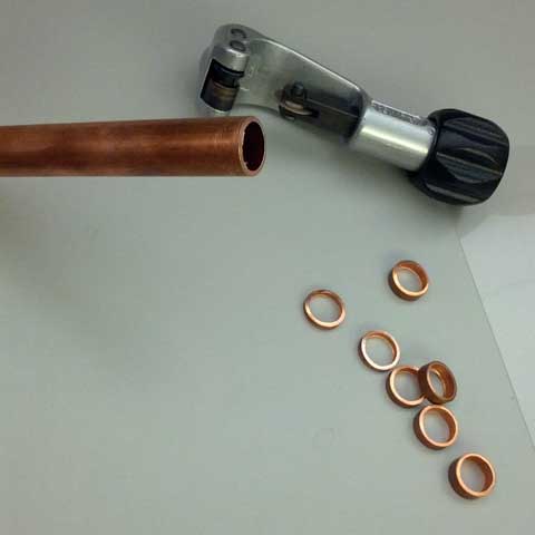 Several rings have been cut from the length of copper pipe and lie on the table next to it, the pipe cutter, and the transparency sheet.